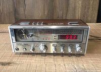 Front view showing analog quartz clock, analog and digital tuning indicators, orange LED frequency display, and control knobs on silver panel with woodgrain top.