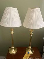 Two brass table lamps side by side on a wooden surface with white pleated lampshades, about 30 inches tall each.