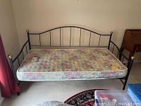 Full view of black aluminum daybed frame with mattress, showing floral pattern mattress and brass ball finials on corners.