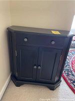 Black finished end table positioned against a corner wall, showing front view of the single drawer and double door cabinet with round knobs. The table size proportions and design are clearly visible with carpet floor surrounding.