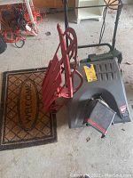 Photo showing Craftsman snowblower, red Radio Flyer sled, and welcome mat on garage floor with cords and workbench in background.