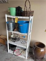 White plastic shelving unit with four shelves holding various metal tubs, baskets, and sports equipment visible beneath. Situated against a beige wall on garage concrete floor.