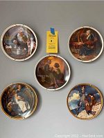 Five Norman Rockwell collectible plates arranged on a grey background, displaying a variety of classic Americana scenes from the artist.