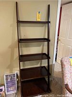 Front view of the dark wood ladder-style shelving unit with five shelves, placed against a wall with a chair and other furniture nearby.