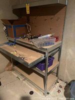 Front view of metal tool bench with pegboard and various tools and supplies on top shelf and work surface.