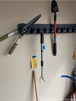 Photo showing garden clippers, three-prong cultivator, and small shovel hanging on a wall rack inside garage.