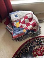 Stacked bedding items on carpet near window, showing twin-sized comforter, quilt, and blanket with patriotic star and checkered patterns
