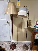 Photo showing two standing lamps side by side near a door and a cabinet, one with a wooden pole and beige shade, the other with a twisted metal pole and striped shade.