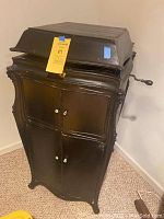 Front angle view of the closed Victrola wooden cabinet with visible crank handle and decorative moldings. Yellow MaxSold tag attached.