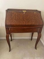 Closed front view of vintage dark wood roll-top secretary desk with curved legs and caster wheels.