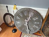 View of the metal wall clock showing large Roman numerals and decorative clock hands, placed on the floor leaning against a wall