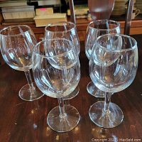 Five clear wine glasses displayed on wooden surface with background showing dining ware and books