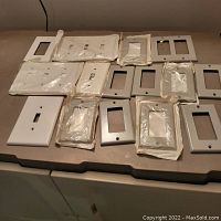 Fifteen white light switch and outlet cover plates arranged on a table, some in original plastic packaging, showing assorted sizes and configurations.