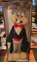 Plush Bugs Bunny dressed in tuxedo with red bow tie and cummerbund inside original display box, holding a black top hat, smiling with large front teeth and big eyes.