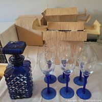 Blue glass decanter with square geometric pattern and stopper beside 4 blue stem wine glasses with etched bowls and twisted stems