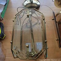 Clear glass indoor canopy light fixture with metal frame placed on a wooden surface. Visible wiring and light socket inside the glass enclosure.