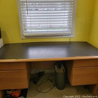 Front view of wooden desk showing top surface and four drawers, placed in front of window with white blinds. Scratches visible on surface.