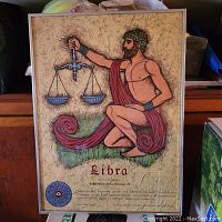 Framed glass poster showing an artistic colored illustration of a man holding Libra scales, with text about the Libra zodiac sign.