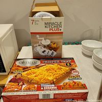 Miracle Kitchen Plus+ boxed 7-in-1 kitchen tool set alongside Gotham Steel Crisper Tray 2-piece set boxes on table with background kitchen items.