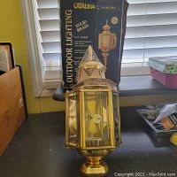 Frontal view of the polished solid brass outdoor light fixture with the product box behind it showing brand and product details.