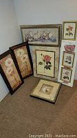 Seven framed floral pictures arranged upright on the floor and leaning against a wall, showing variety in frame styles and flower designs