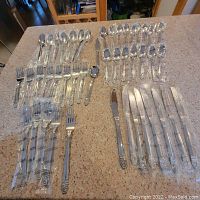 Flatware pieces laid out on countertop, grouped by type: knives, forks, spoons. Most items wrapped in clear plastic packaging. Visible ornate design on handles.