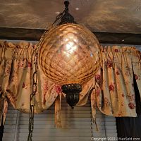 Photo of entire hanging light showing the amber textured globe suspended by a brass chain in front of window with floral curtains.