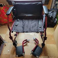 Front view of the lightweight foldable wheelchair with red frame and black fabric seat, along with detachable footrests placed in front.