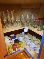 Shelf of assorted champagne flutes, wine glasses with etched designs, clear drinking glasses with red rims, and various coffee mugs including ceramic tiki mugs and branded mugs.