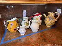 Six assorted porcelain pitchers shown on countertop, varying styles and sizes, some floral and colorful designs
