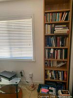 Tall wooden bookshelf filled with many books in different sizes and colors. Books arranged on shelves from top to bottom with some titles visible, a box on the floor with additional books or magazines.