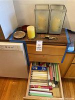 Assorted taper, votive, and pillar candles in an open drawer with two glass candleholders on a countertop next to metal trivets.