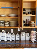Kitchen open cabinets showing stacked stoneware plates and bowls on left side, with assorted black and blue mugs on the right side shelves.