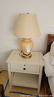 Photo of the Ethan Allen nightstand with cream finish, one drawer, pull-out writing surface, and bottom shelf, lamp placed on top.