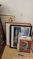7 framed or mounted art prints and photographs leaning against a wall and a dresser showing a variety of frame styles and sizes.