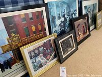 Seven framed reproductions of various art prints arranged on floor leaning against wall and furniture. Varied frame styles and sizes.