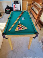 Miniature pool table full view showing wooden frame and legs, two pool cues, full set of balls in rack, cue chalk on playing surface