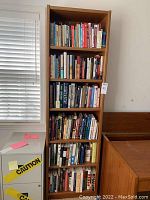 Front view of one tall, narrow oak veneer bookcase filled with books on all shelves, placed near a window.