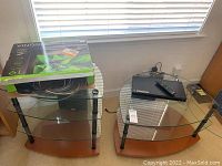 Photo of two glass shelf media stands with metal legs and wooden bases, assorted cords in box on top of left stand, Sony Blu-ray player and remote on right stand near window.