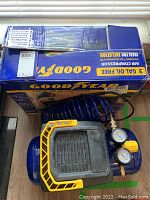Photo showing boxed Goodyear 3 gallon oil-free air compressor with blue coiled air hose and black and yellow protective casing on the unit.