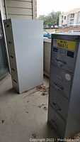 Photo of two gray vertical metal file cabinets on a balcony or outdoor covered space, showing size and condition.