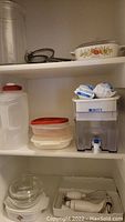 Shelf showing plastic food containers with red lids, Brita water filter dispenser with two filters on top, glass mixing bowl with lid and handheld wand mixer with beaters.