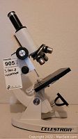 White Celestron microscope with multiple lenses and adjustment knobs on a beige background.