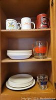 Photo showing three distinct ceramic mugs on top shelf, stack of white bowls on middle shelf with tall glass measuring cup, and white plates on bottom shelf with smaller glass measuring cup.