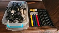 Plastic container holding multiple coiled power cords and cables in black and white colors. Next to it is a black plastic case containing hand tools and a small bottle of solution.