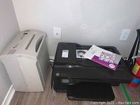 Combined view of Fellowes Powershred PS 60CC shredder and HP Officejet 7612 printer placed side by side on wooden floor near wall electrical outlets.