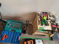Wide view showing wooden nightstand missing one drawer, multiple tool sets and loose tools on and around it including blue and green cases of sockets and hand tools.