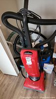 Red Dirt Devil upright vacuum cleaner with attached hose and cords, standing on wood floor.