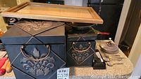 Two decorative black metal boxes with embossed floral designs and bronze-colored handles, wooden picture frame, metal bowl, metal utensils, oil lamp, and a sensor watch on the table.