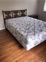 Full view of bed including wood headboard carved with symmetrical scroll motif, metal frame base, and floral mattress on hardwood floor.